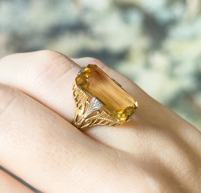 Angled top view shown on a finger, the pale orange-yellow stone lying diagonally across the knuckle so its long facets and both filigree shoulders are visible.