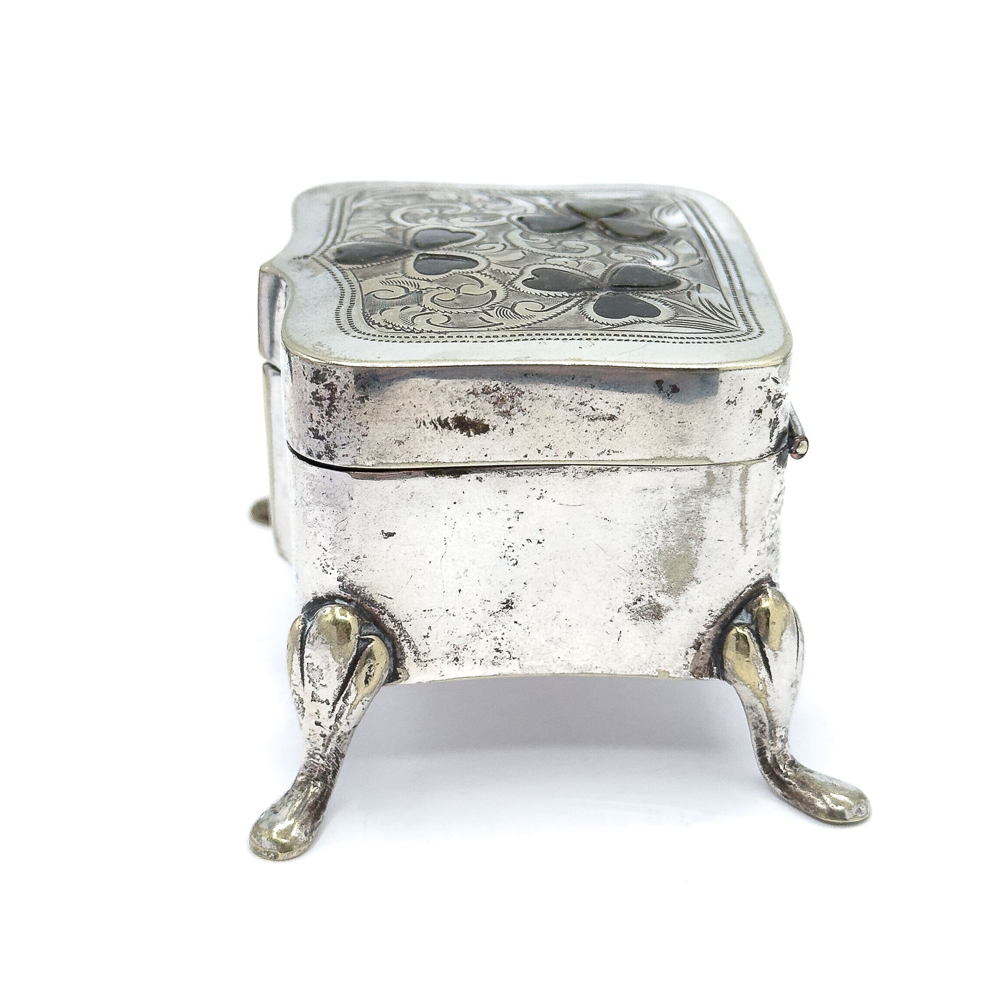End view from the narrow short side, showing the box's depth, the seam where the lid meets the body, a small catch at the right edge, and two teardrop feet splaying forward.
