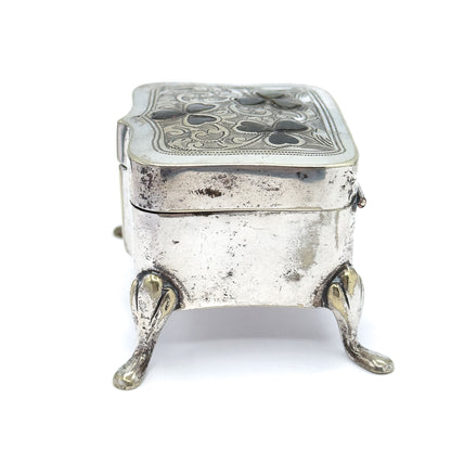 End view from the narrow short side, showing the box's depth, the seam where the lid meets the body, a small catch at the right edge, and two teardrop feet splaying forward.
