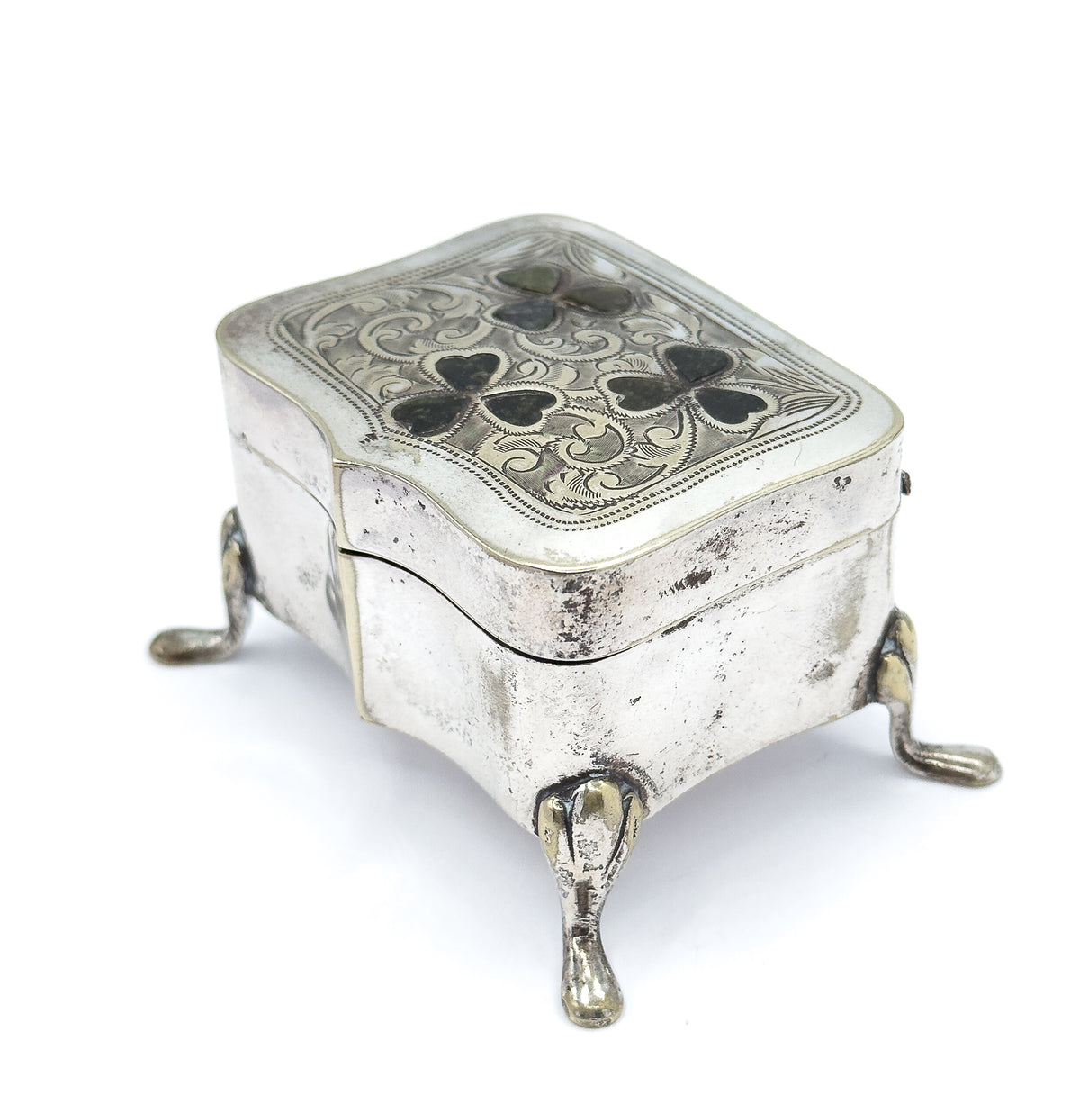 Angled from a front corner with the lid catching the light, so the scrollwork and dark clover insets read across the whole face while three teardrop feet and the hinged seam show below.