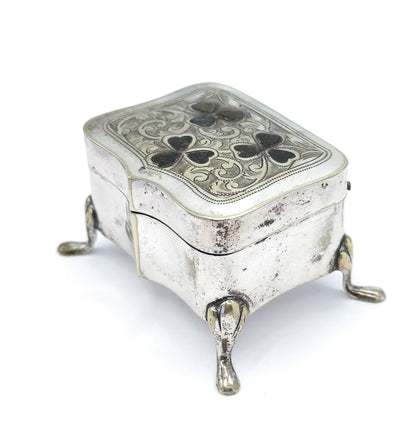 Angled from a front corner with the lid catching the light, so the scrollwork and dark clover insets read across the whole face while three teardrop feet and the hinged seam show below.