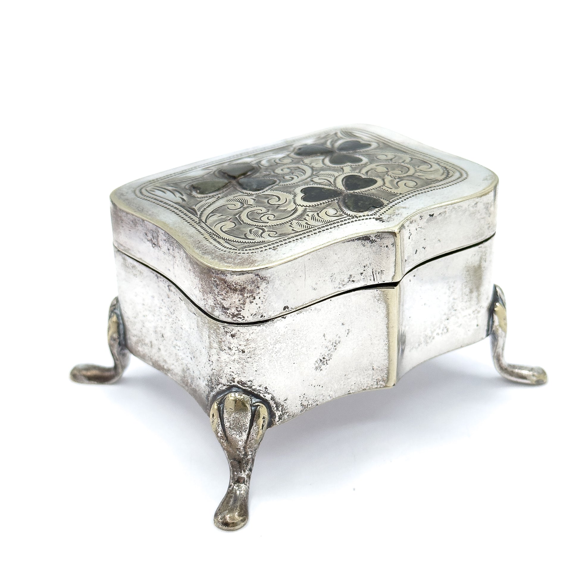 Low three-quarter view from a corner, the serpentine sides rising to the engraved lid with its clover insets while three teardrop feet splay out beneath the shaped base.