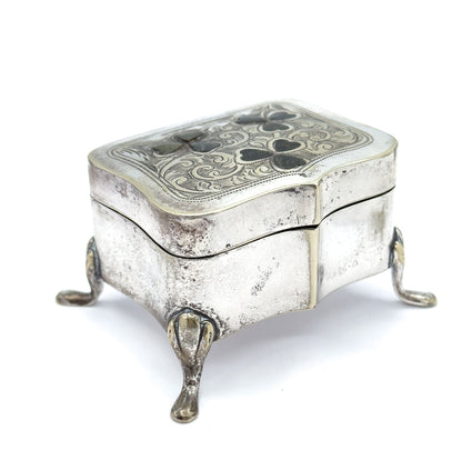 Low three-quarter view from a corner, the serpentine sides rising to the engraved lid with its clover insets while three teardrop feet splay out beneath the shaped base.