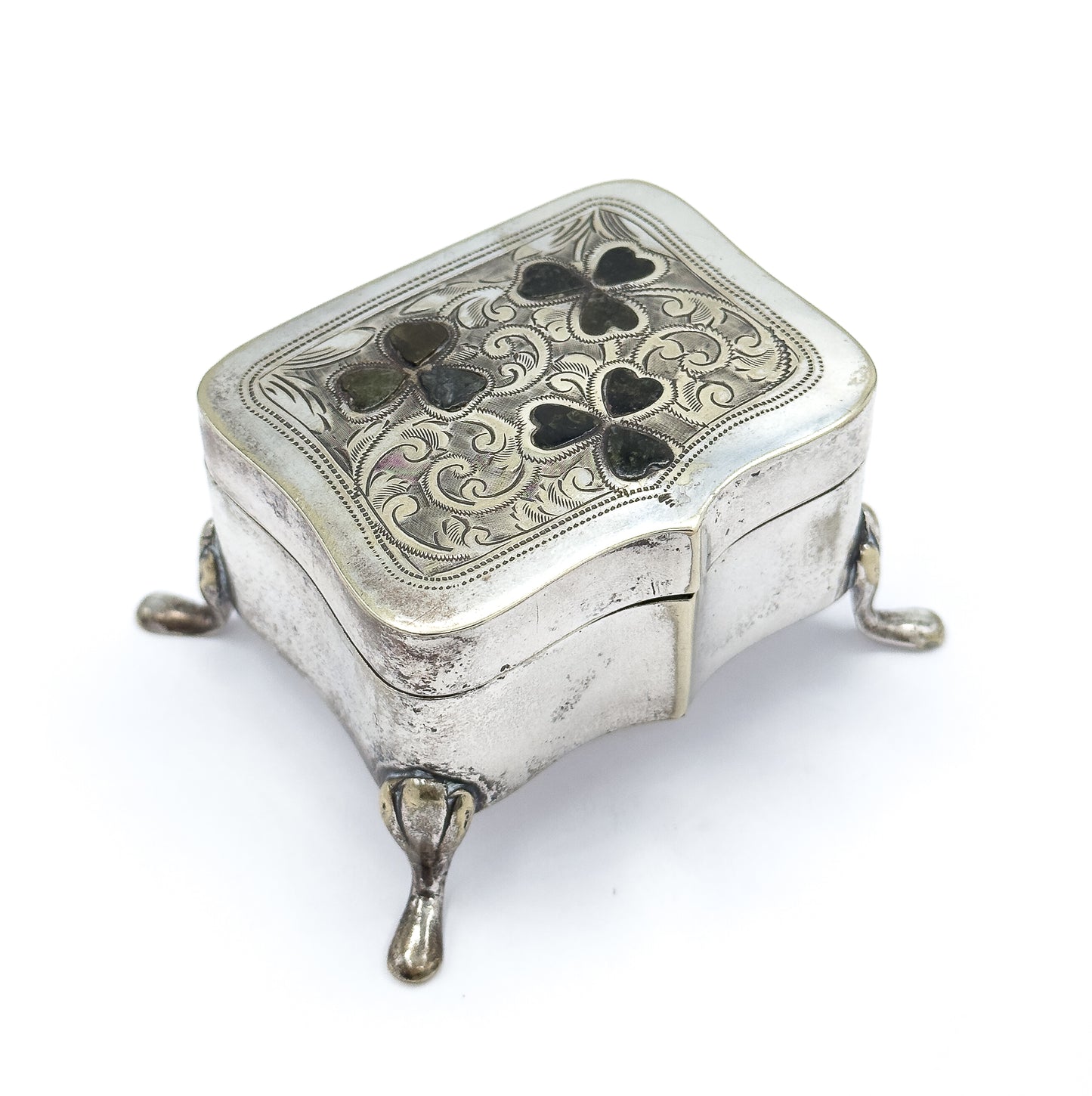 Tilted from above and slightly behind one corner, the lid's seam and hinge line run along the far side while the engraved face with its three green clovers and the teardrop feet stay in frame.
