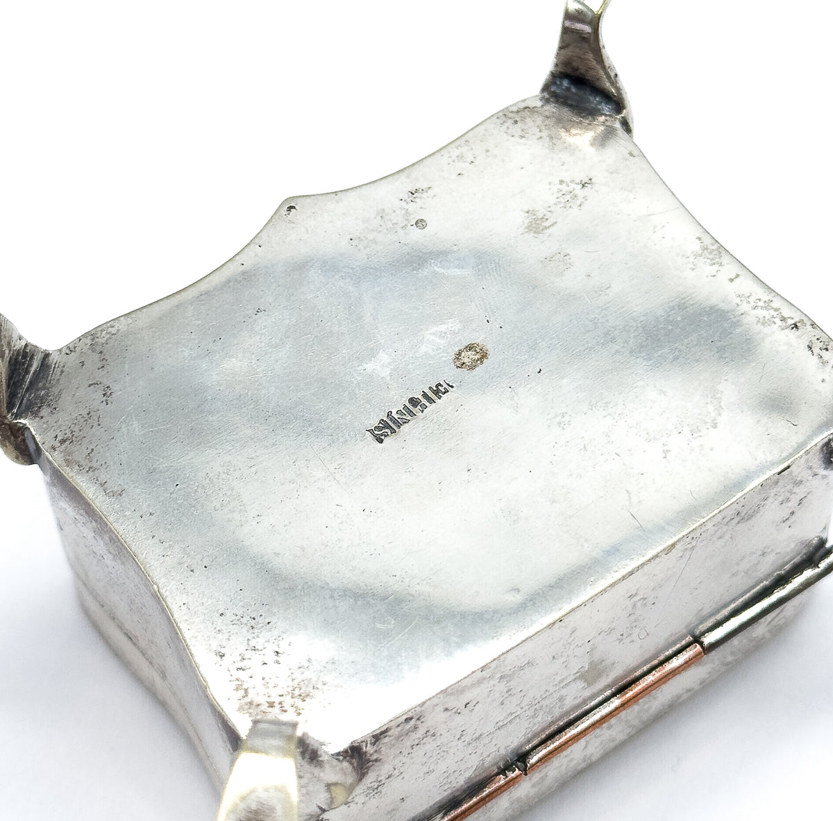 The underside of the box tipped up to show its shaped base with a small stamped mark at the centre, the hinge and two little feet visible at the edges.
