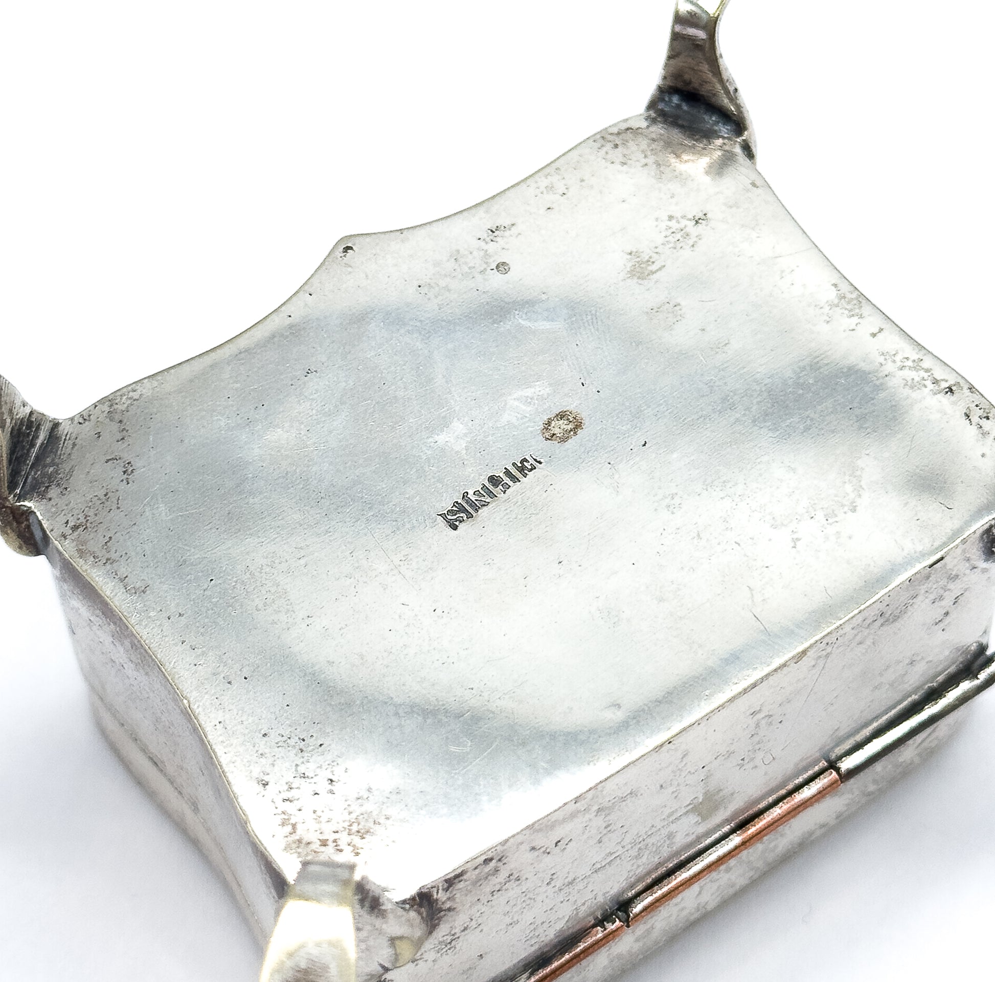 The underside of the box tipped up to show its shaped base with a small stamped mark at the centre, the hinge and two little feet visible at the edges.