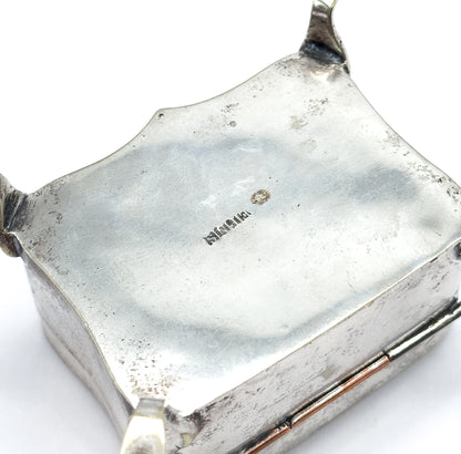 The underside of the box tipped up to show its shaped base with a small stamped mark at the centre, the hinge and two little feet visible at the edges.