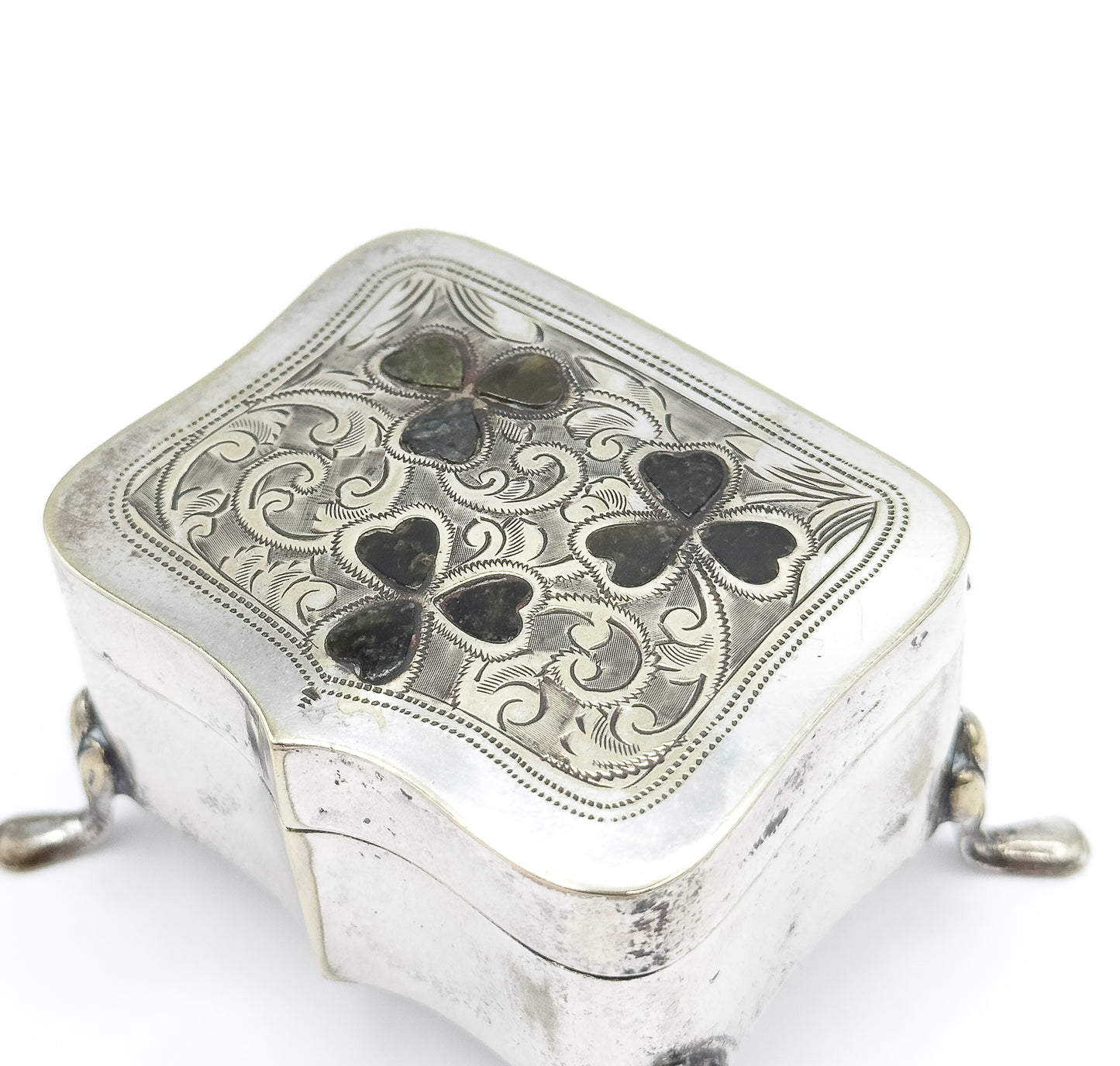 Close overhead view of the closed lid, its beaded border framing dense engraved scrolls around the three green marble clovers, with the hinge line and two feet showing at the sides.