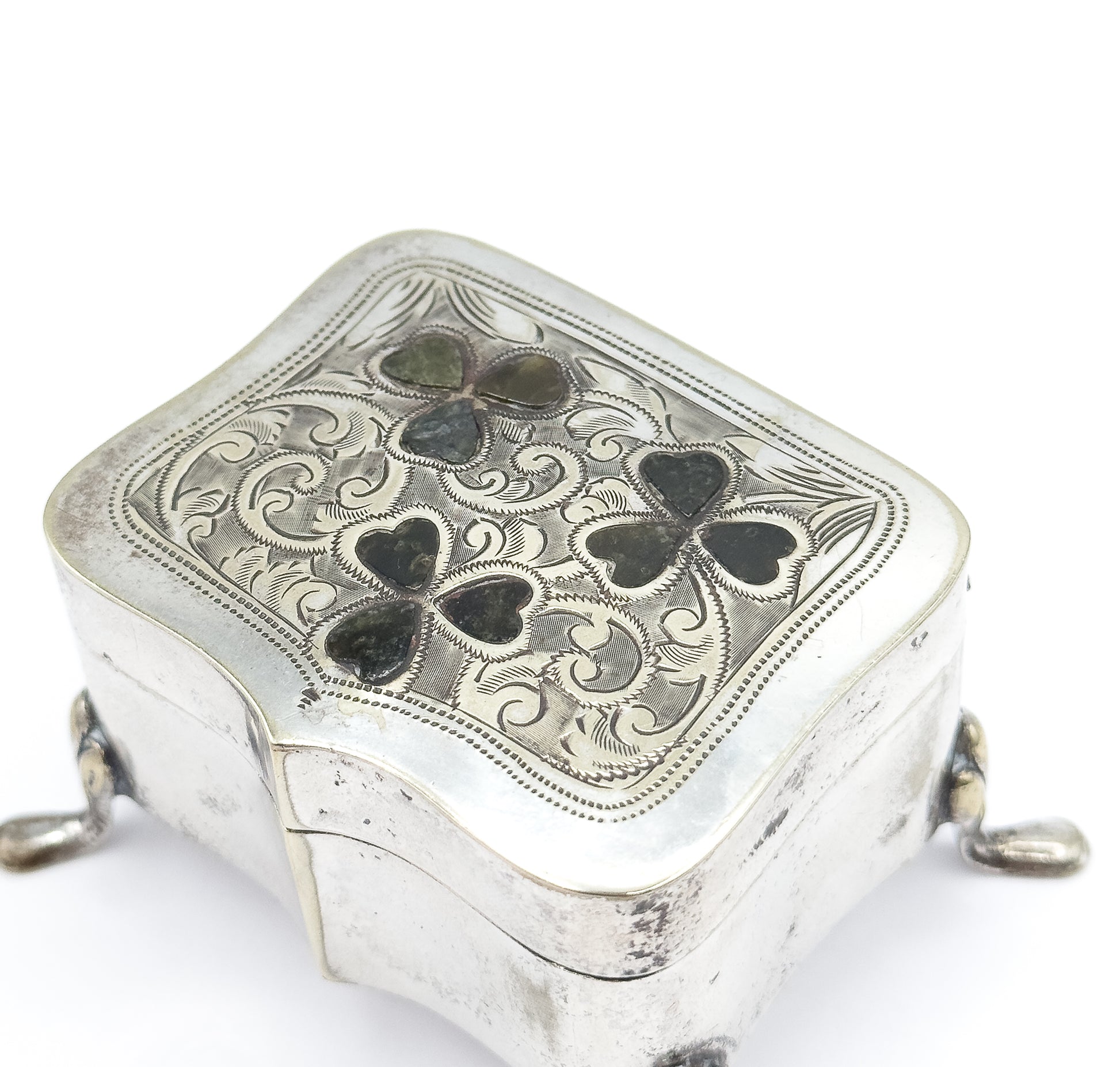 Close overhead view of the closed lid, its beaded border framing dense engraved scrolls around the three green marble clovers, with the hinge line and two feet showing at the sides.