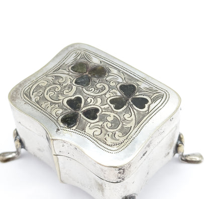 Close overhead view of the closed lid, its beaded border framing dense engraved scrolls around the three green marble clovers, with the hinge line and two feet showing at the sides.