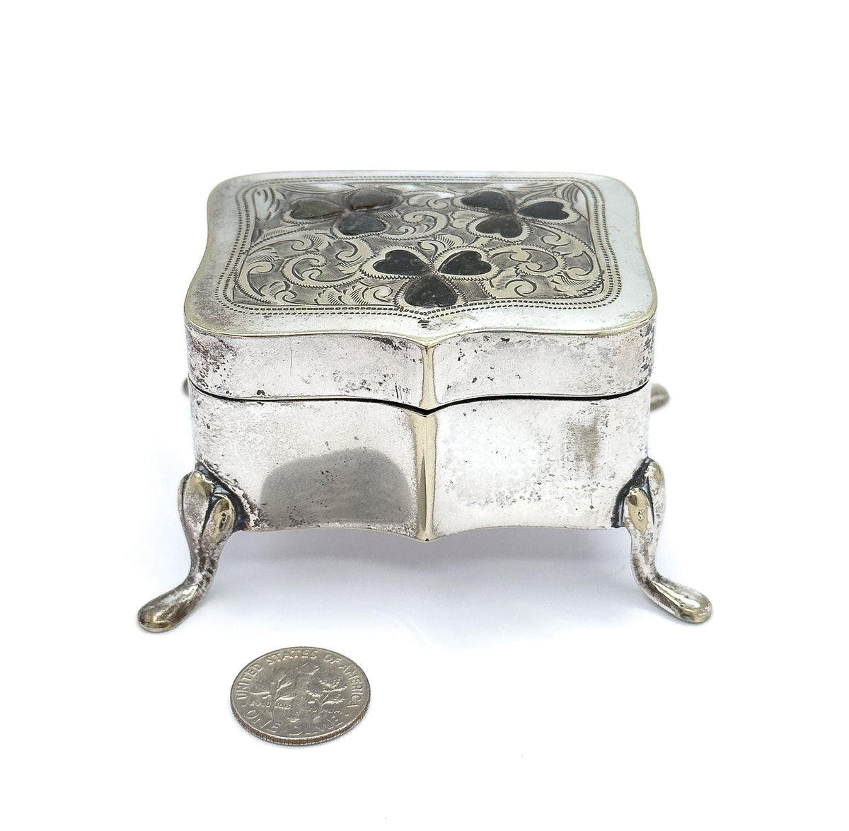 Straight-on front view of the closed box on its four splayed feet, with a coin placed in front to convey its small size and the engraved clover lid seen from the front edge.