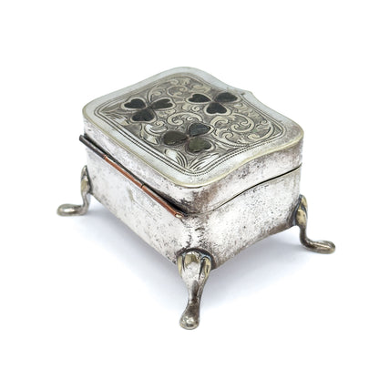 Lower three-quarter view from the hinged back edge, where the hinge barrel runs along the side, the plain engraved body sits deep above two splayed teardrop feet, and the shaped lid overhangs.