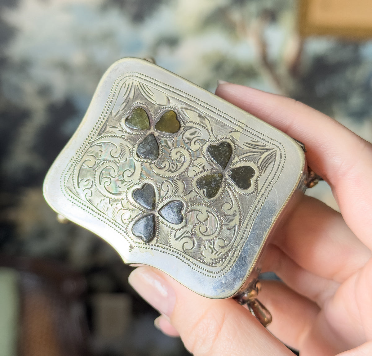 Edwardian Connemara marble clover trinket box in silver plate, held in a hand that shows its palm-filling size, with three green marble clovers set into the hand-engraved lid.