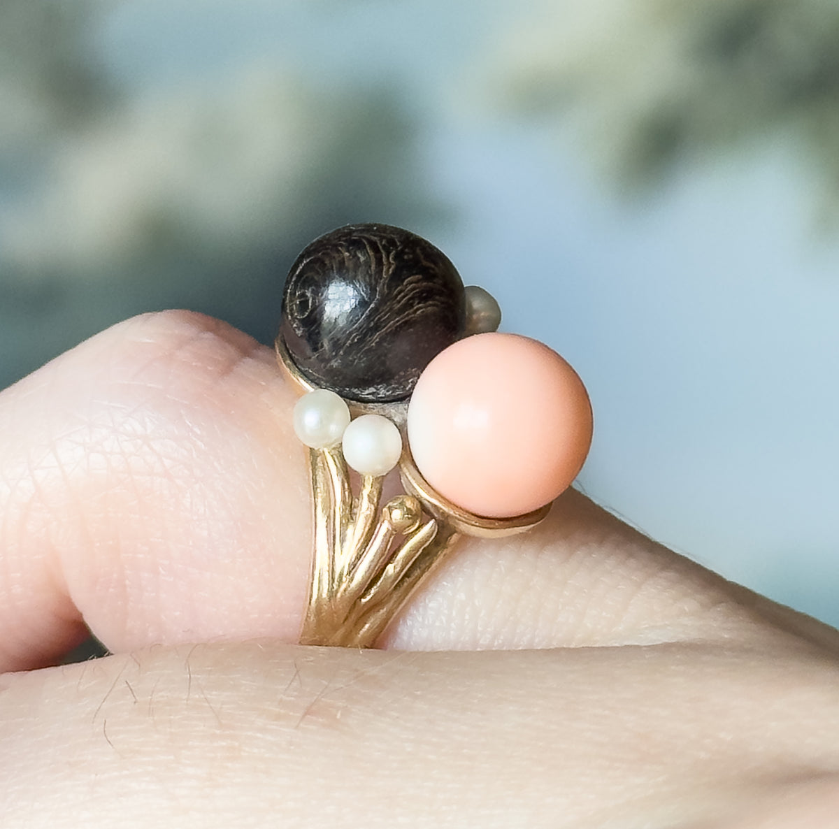 Close side view shown on a finger, the peachy pink and dark coral orbs sitting shoulder to shoulder above three seed pearls and the yellow gold shank that splays into slender prongs.