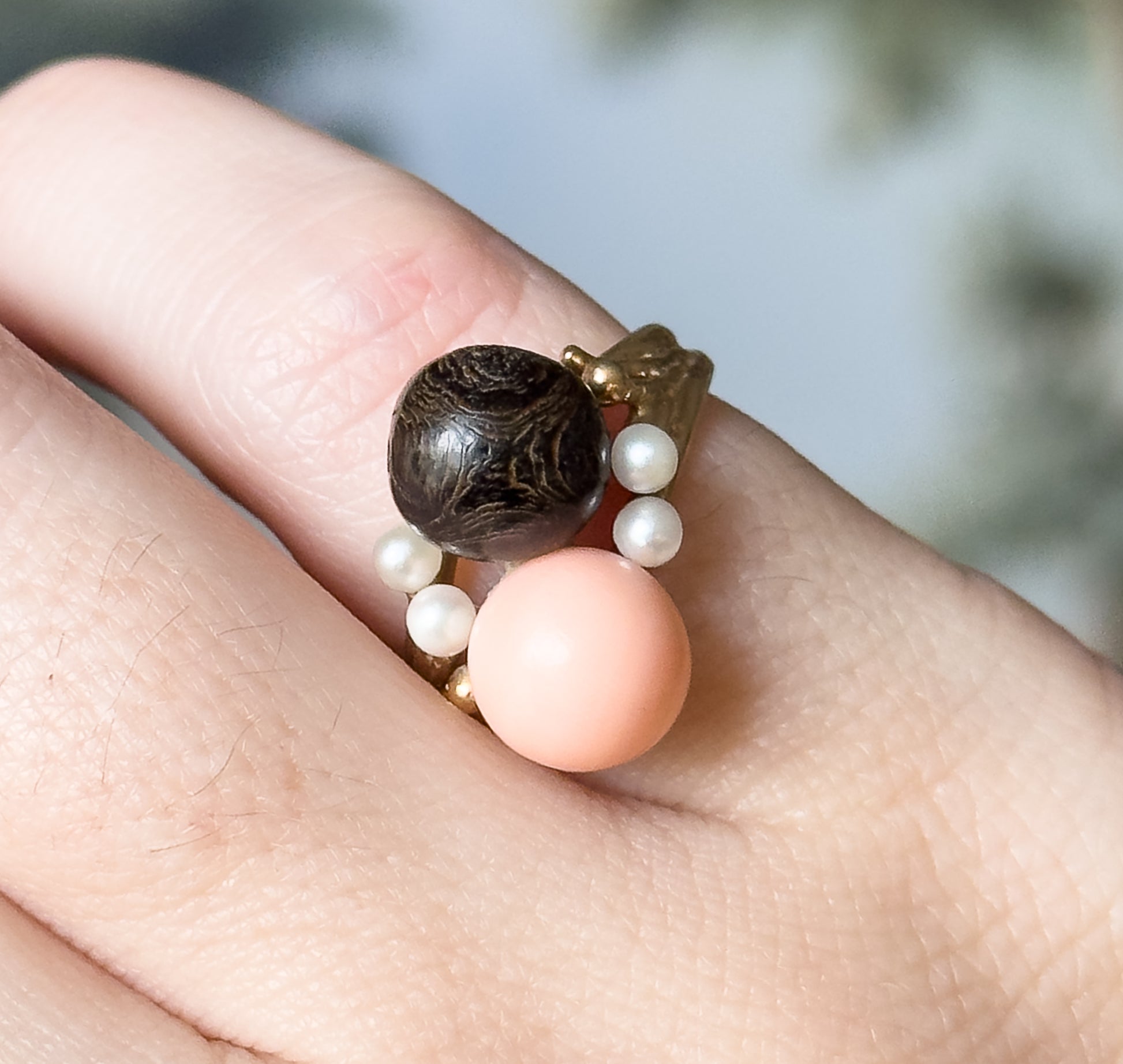Overhead view shown on a finger, looking straight down onto both coral orbs with four seed pearls ringing them, the finger giving a clear sense of the ring's small scale.