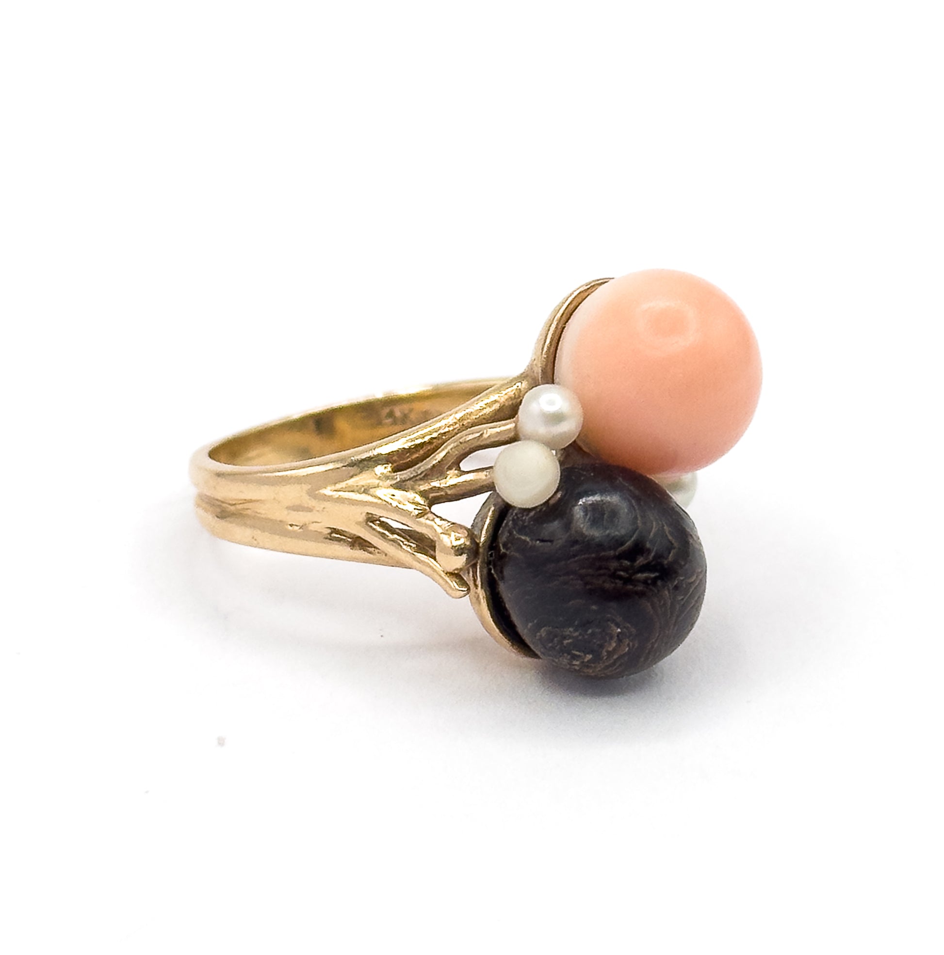 Angled side view against white showing the textured branch-like shoulders rising from the split shank to hold the peachy pink orb above and the dark coral orb below, with seed pearls between.
