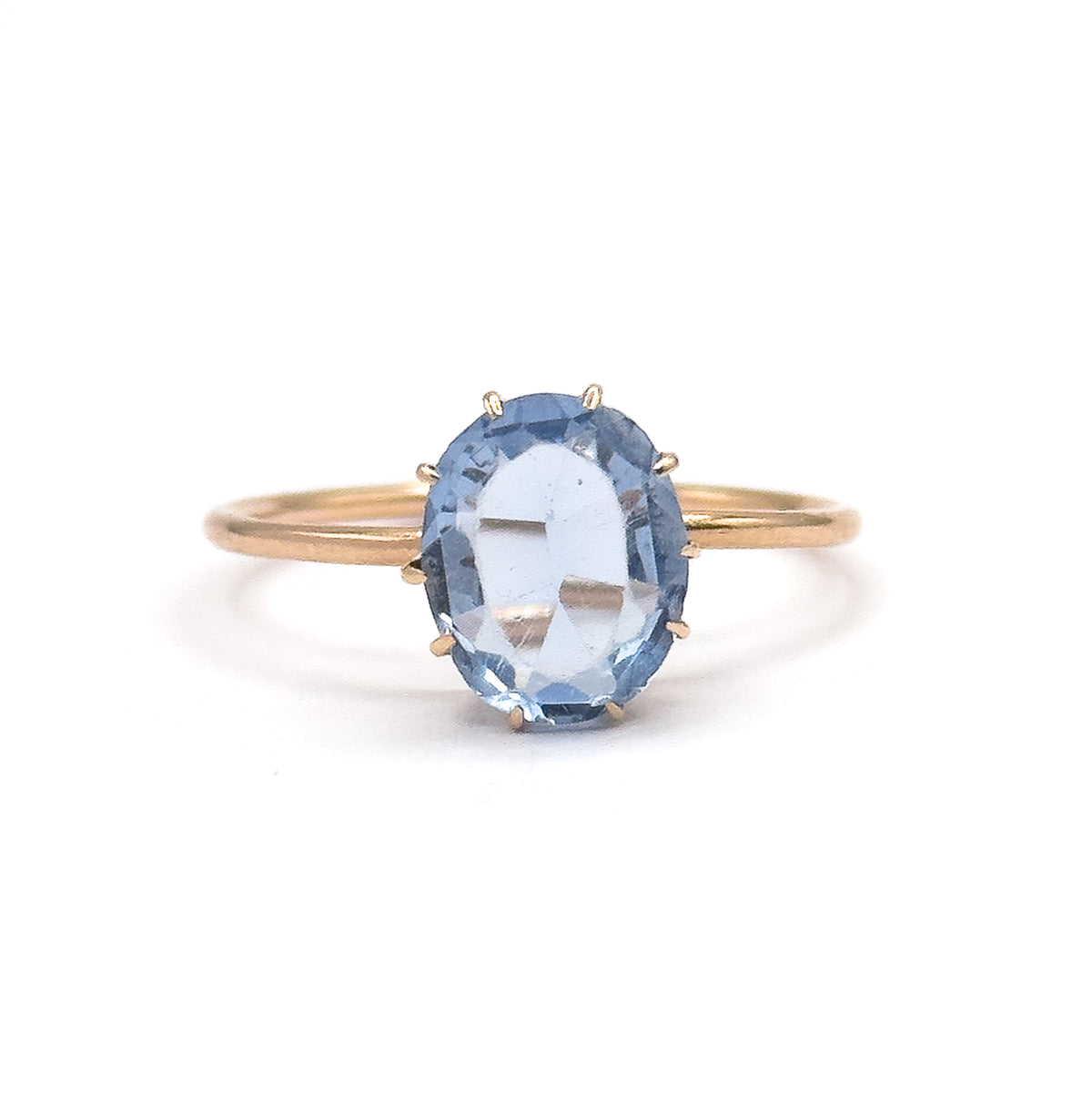 Straight-on view against plain white, the whole slim yellow gold shank forming a complete circle behind the oval purplish blue glass stone held in its prongs at the centre.