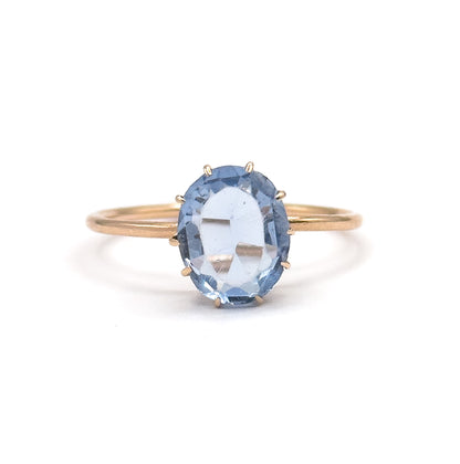 Straight-on view against plain white, the whole slim yellow gold shank forming a complete circle behind the oval purplish blue glass stone held in its prongs at the centre.