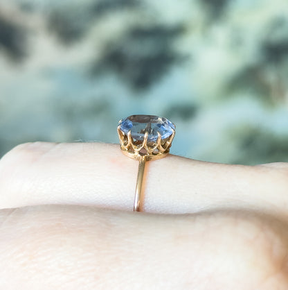 Side-on view close to the finger, showing the open scalloped gallery, the prongs gripping the purplish blue glass stone, and the slender yellow gold shank below.