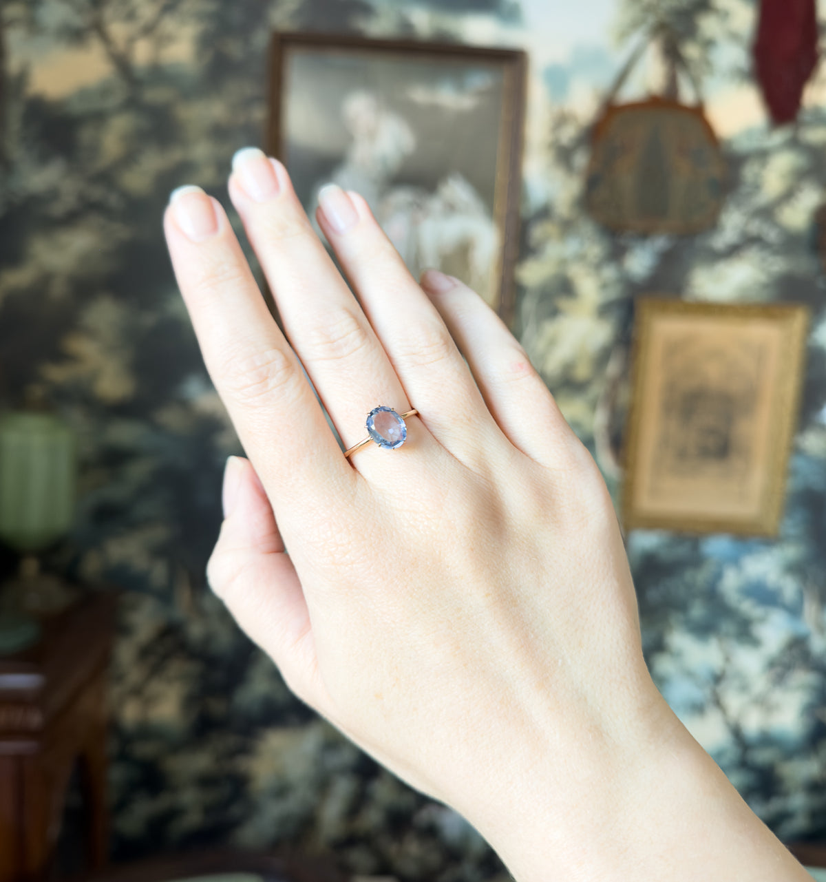 Full view of the hand raised against a leafy patterned backdrop, the small ring sitting low on the ring finger so its modest size against the whole hand is clear.