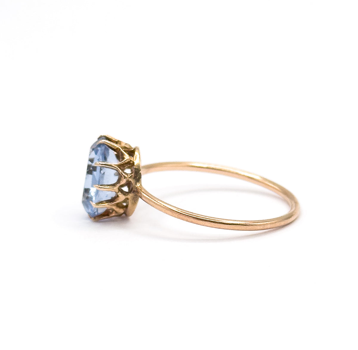 Close side view filling the frame, the slender yellow gold shank sweeping back from a scalloped gallery whose long prongs grip the purplish blue glass stone in profile.
