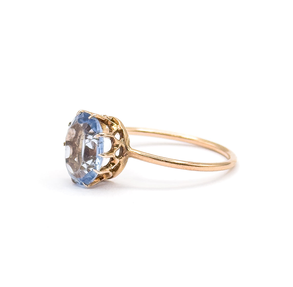 Side view from the opposite direction, the oval glass stone tilted toward the camera so its pale purplish blue table and faceted edge show above the pierced gallery and thin yellow gold shank.