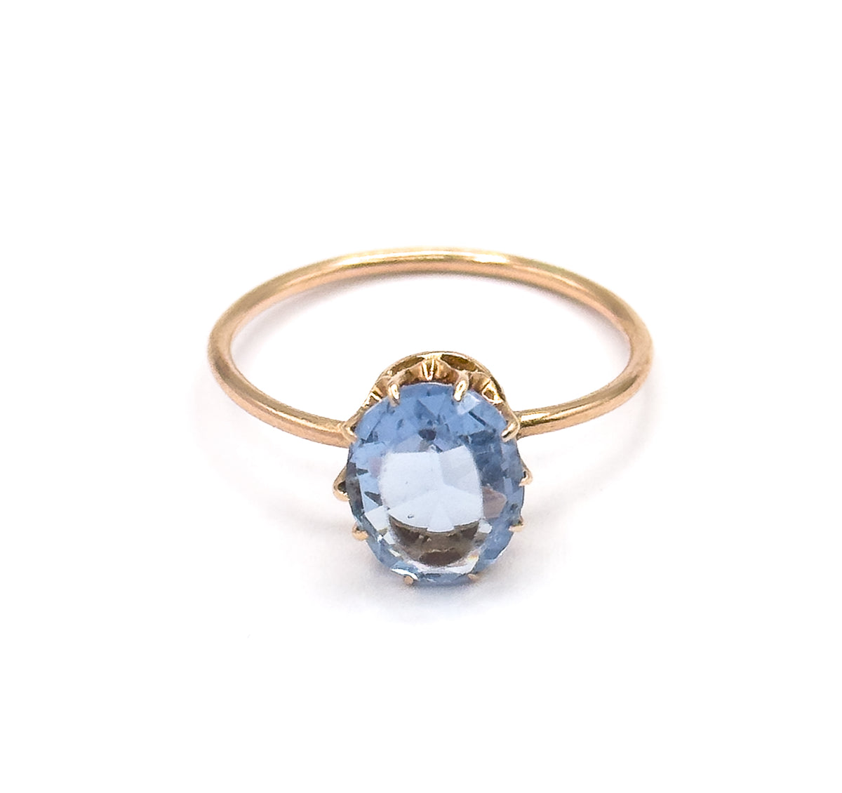 Front-facing view with the ring upright, the oval purplish blue glass stone squarely to the camera showing its facets and the four prong tips, the slim yellow gold shank curving behind.