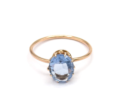 Front-facing view with the ring upright, the oval purplish blue glass stone squarely to the camera showing its facets and the four prong tips, the slim yellow gold shank curving behind.