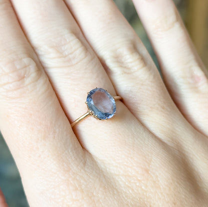 A 14K yellow gold solitaire ring with an oval purplish blue glass stone in a four-prong setting, shown on a finger with a slim shank, giving a sense of its petite scale.