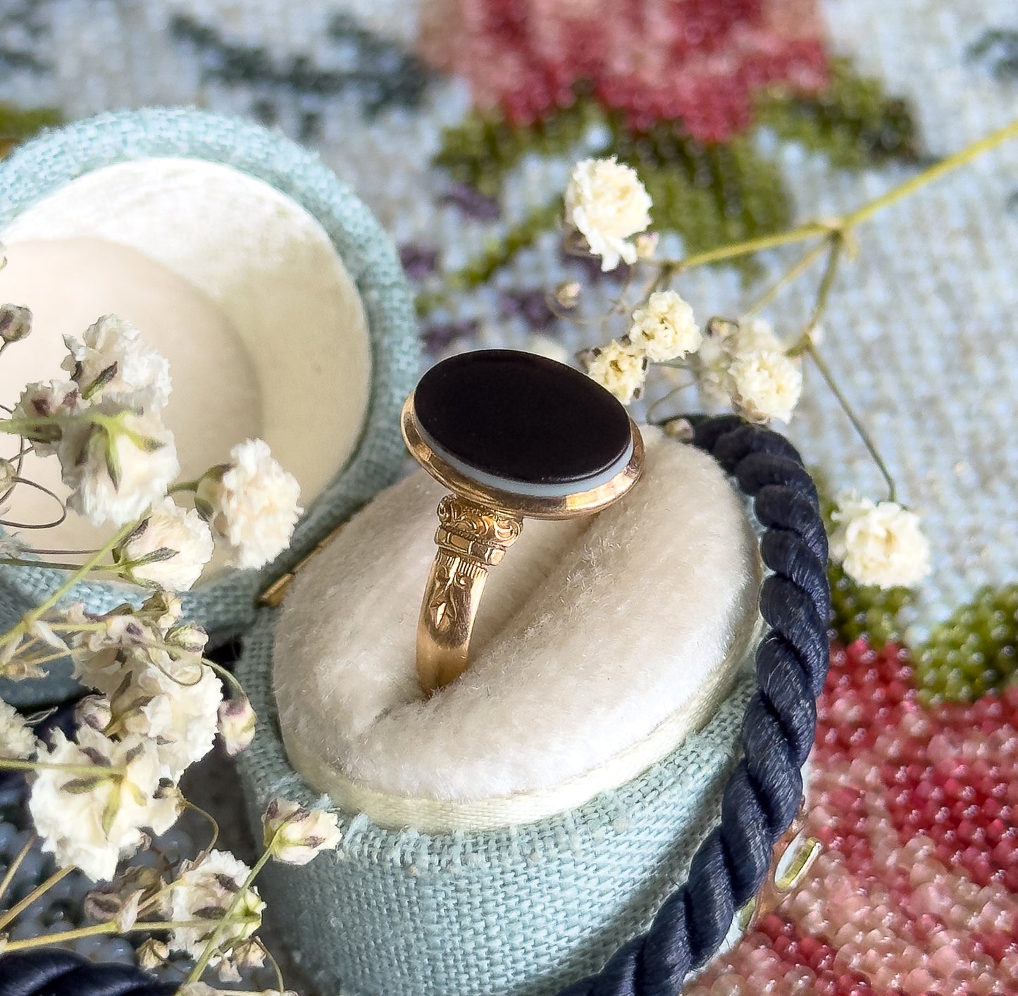 Propped in a pale blue velvet ring box among dried baby's-breath sprigs and a floral tapestry, the black-and-white banded agate and engraved shoulders angled up toward the camera.