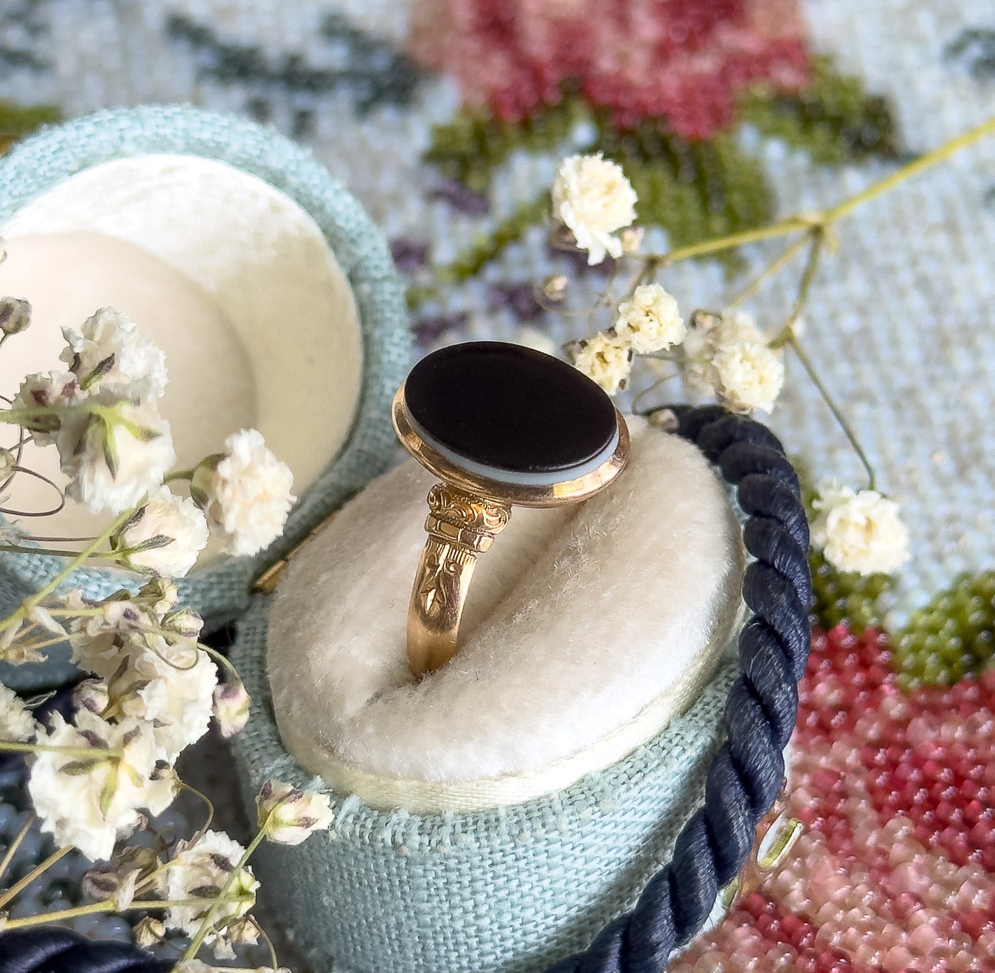 Propped in a pale blue velvet ring box among dried baby's-breath sprigs and a floral tapestry, the black-and-white banded agate and engraved shoulders angled up toward the camera.