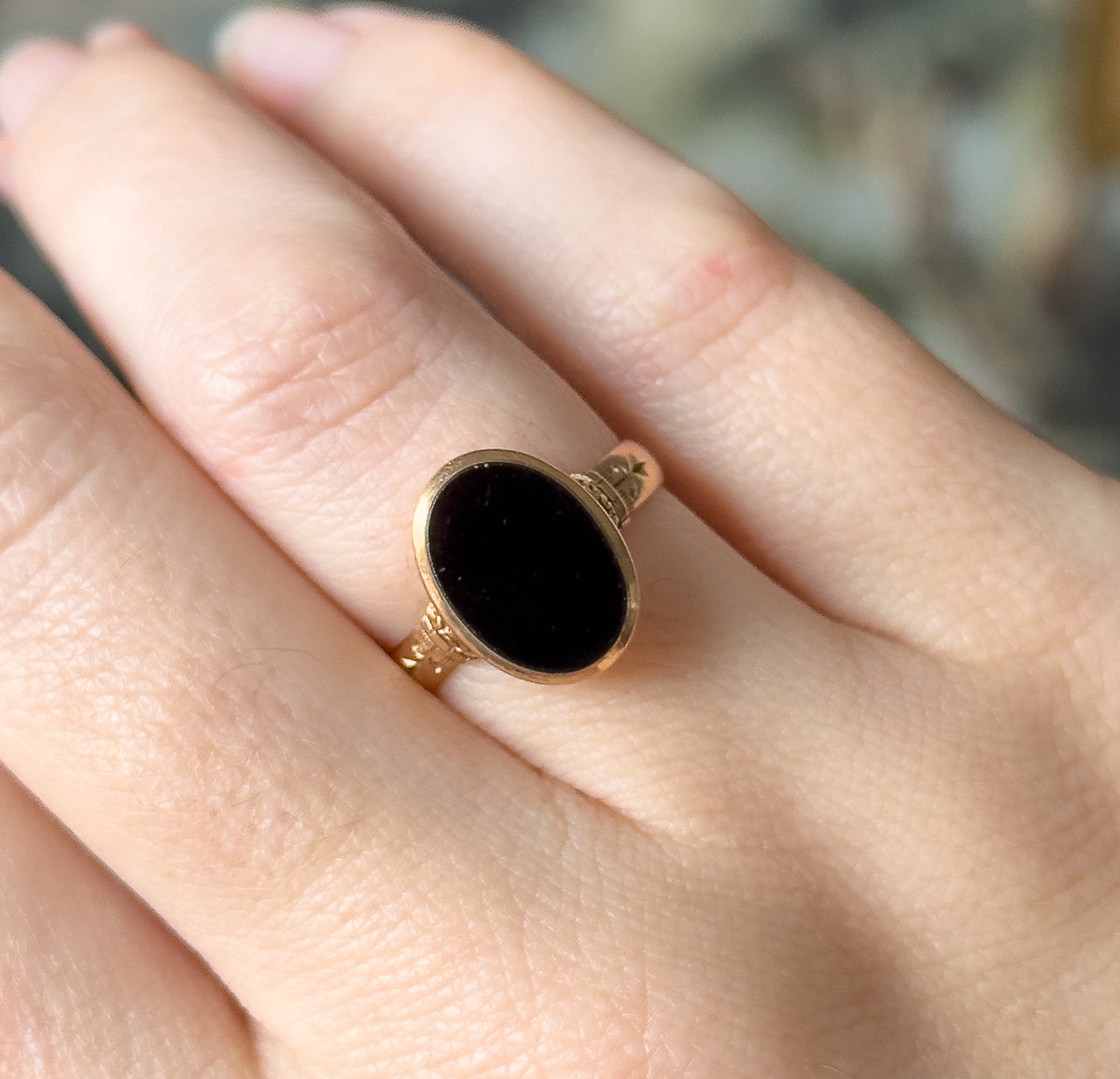 Shown flat-on from directly above on the finger, the whole oval face reading as solid black within its rosy yellow bezel, with the engraved shoulders and narrow shank meeting the skin.
