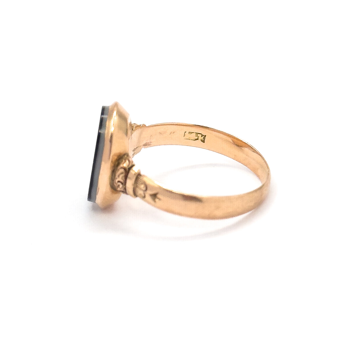 Side view against white with the ring's interior visible, showing a small stamped mark inside the rosy yellow shank, the engraved scrollwork shoulder and the dark stone edge-on at the bezel.