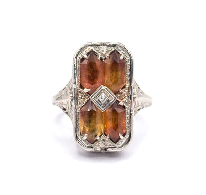 Close overhead view filling the frame with the rectangular face, showing each citrine's long faceted top, the corner prongs gripping them, and the diamond in its square bezel at the centre.
