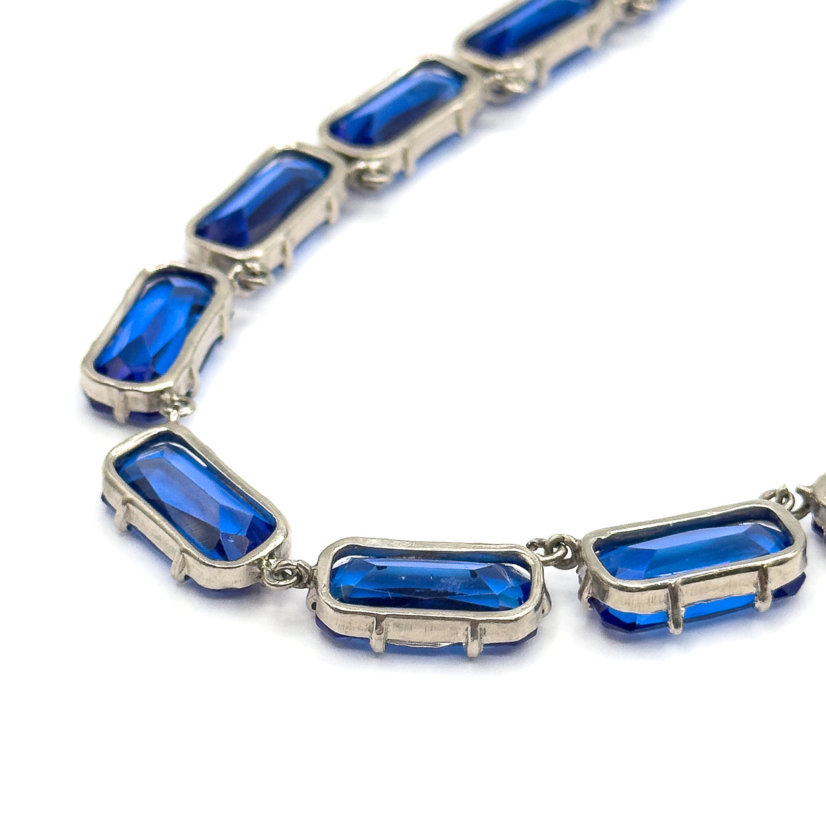 Close-up along a curving run of links, showing the prongs gripping each cobalt blue glass stone and the small jump rings joining one light-coloured metal setting to the next.