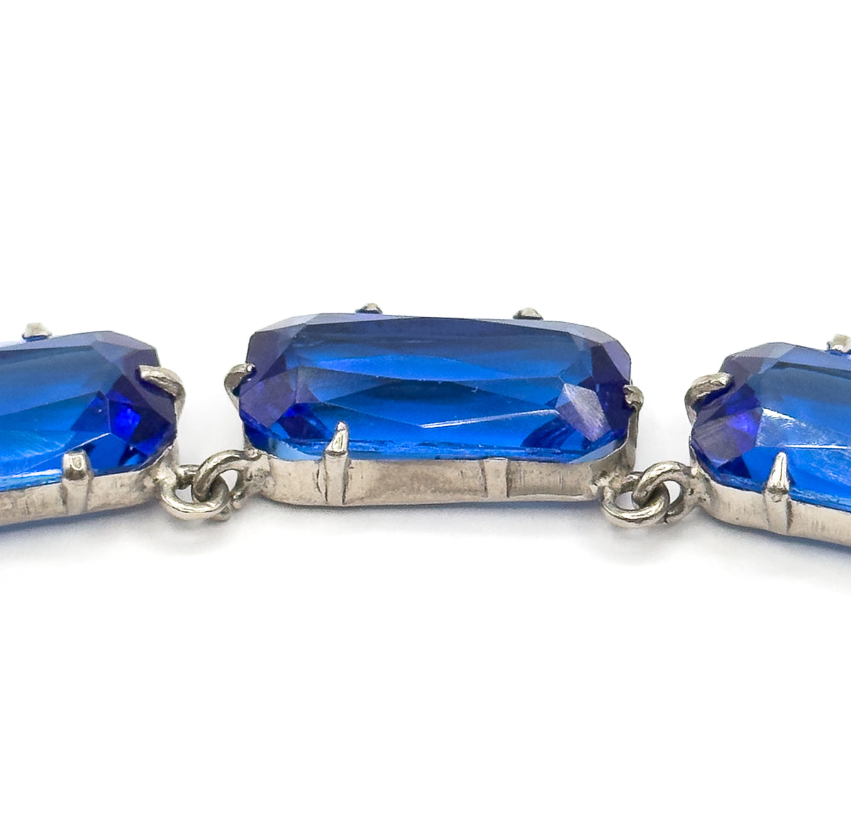 Extreme close-up of three links seen straight on, the facets inside each cobalt blue glass stone catching the light above the smooth metal bezel walls and the paired jump rings between them.