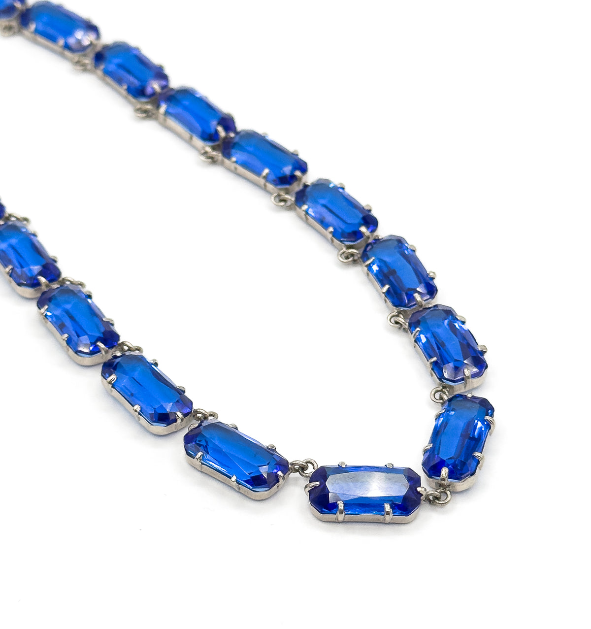 A long diagonal sweep of the necklace against white, the pronged links tapering smaller into the distance so the repeating rhythm of cobalt blue glass stones can be followed end to end.