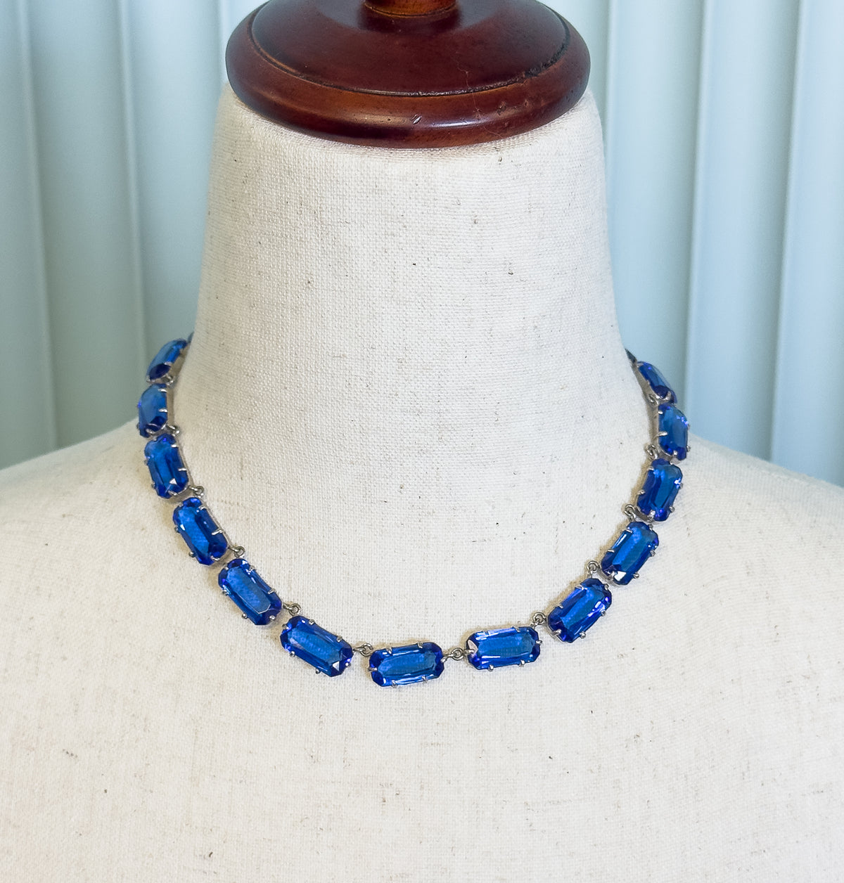 A collar necklace of cobalt blue glass stones in prong-set rectangular links, shown at the neck of a cream fabric display bust.