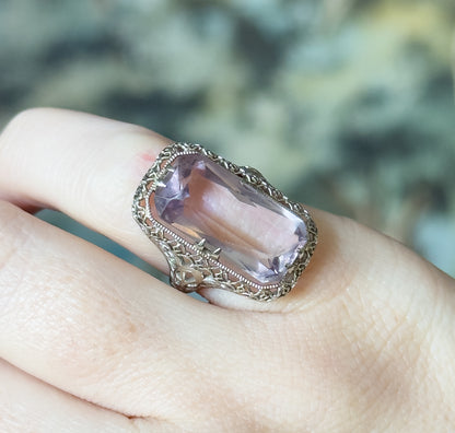 Closer view shown on a finger from above, the pale purple stone's long facets catching light between double prongs, with the lace-like filigree gallery visible along the lower edge of the face.
