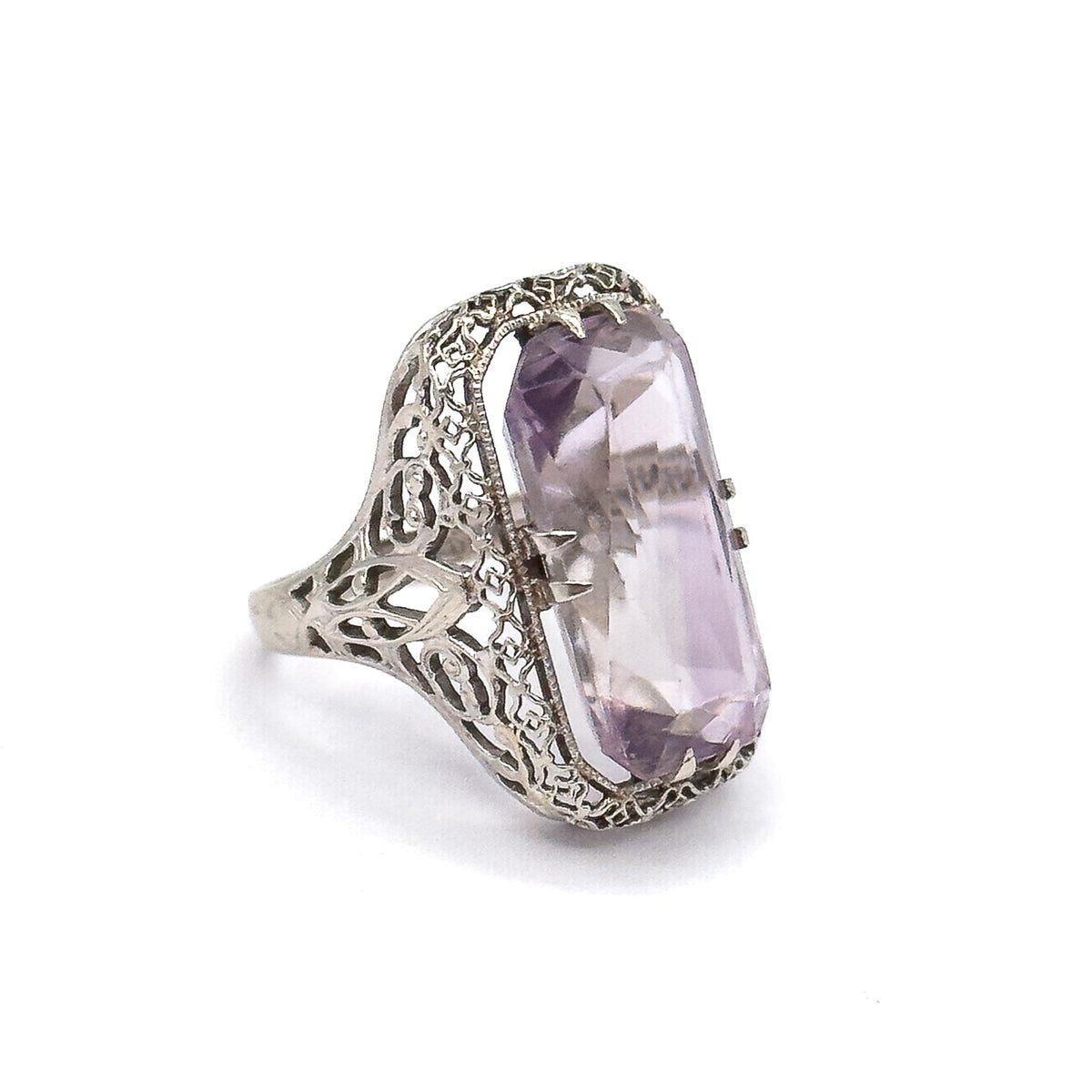 Off the hand against white, the ring stands upright at a three-quarter angle, showing the pierced filigree side gallery, double prongs gripping the pale purple stone, and the tapering shank.