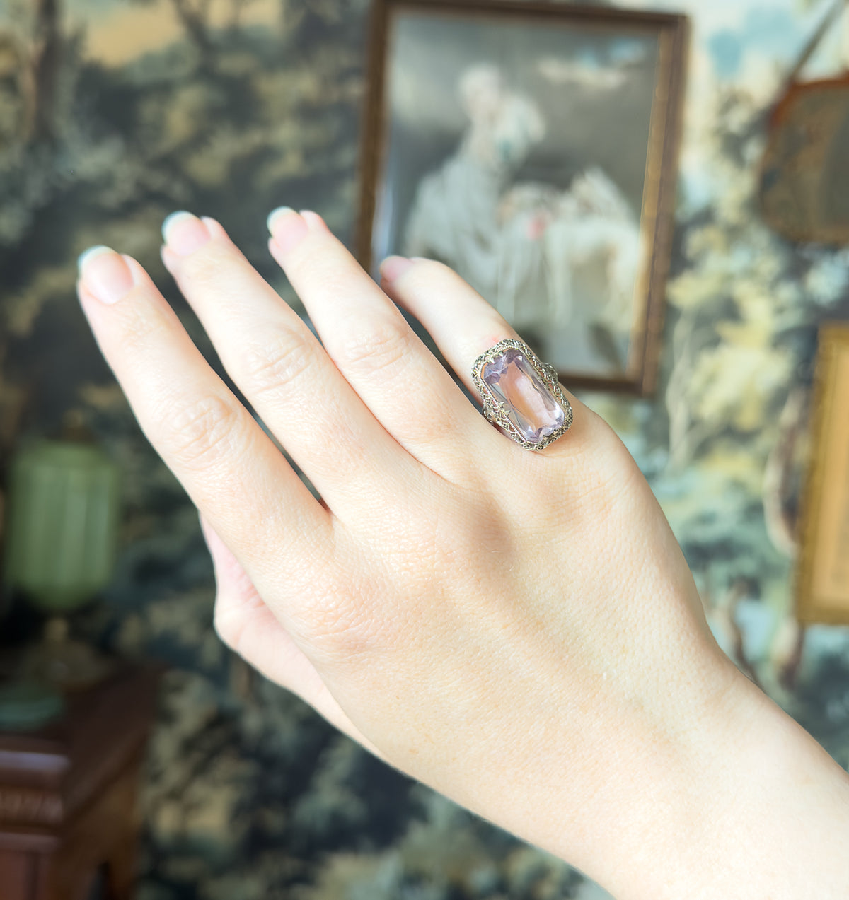 Art Deco amethyst filigree ring in 14K white gold, shown on a finger of a raised hand, the elongated pale purple stone set lengthwise in a milgrained face against a soft tapestry backdrop.