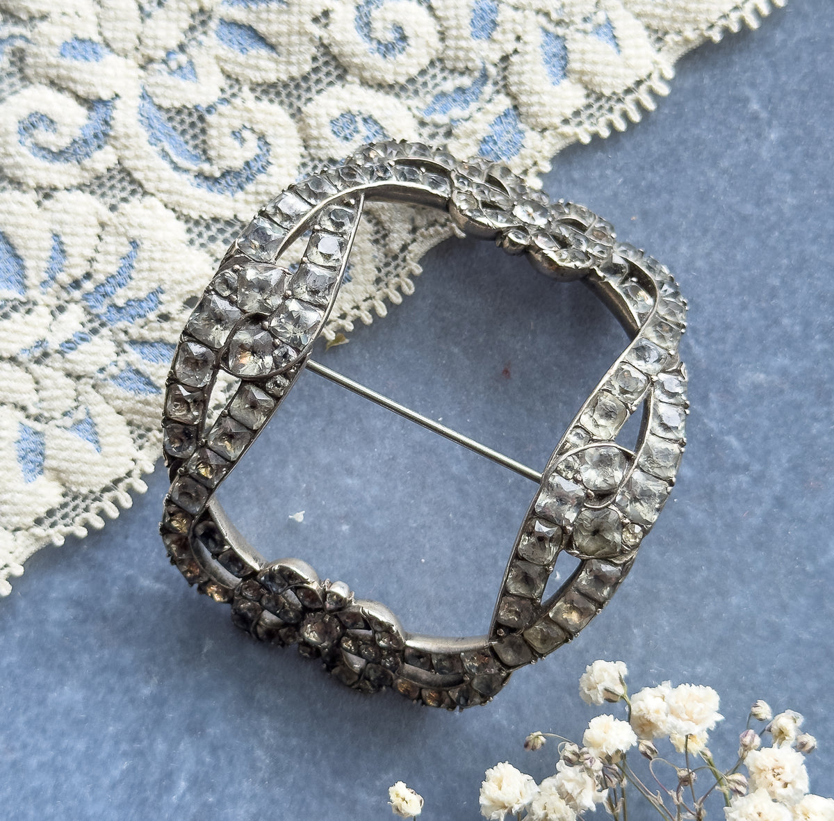 Georgian paste bow buckle brooch in sterling silver, shown face-up on a blue-grey surface beside cream lace and tiny white flowers, its open oval frame set with rows of colourless paste stones.