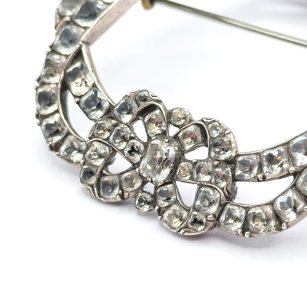 Front macro of the buckle's sweeping curved band, its unbroken line of square paste stones in silver collets curling toward the central bow knot, with the pin stem crossing white above.