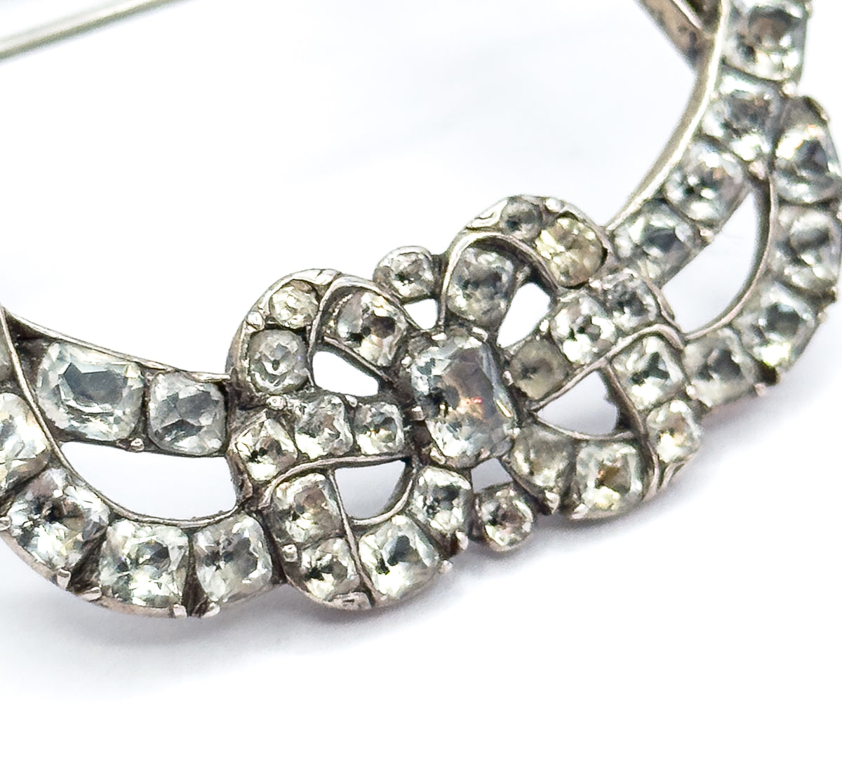 Macro across the lower half of the buckle, where two concentric curved rows of square paste stones in silver collets frame the knotted bow, the pin stem sweeping across white at top left.