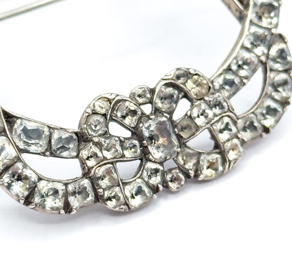 Macro across the lower half of the buckle, where two concentric curved rows of square paste stones in silver collets frame the knotted bow, the pin stem sweeping across white at top left.