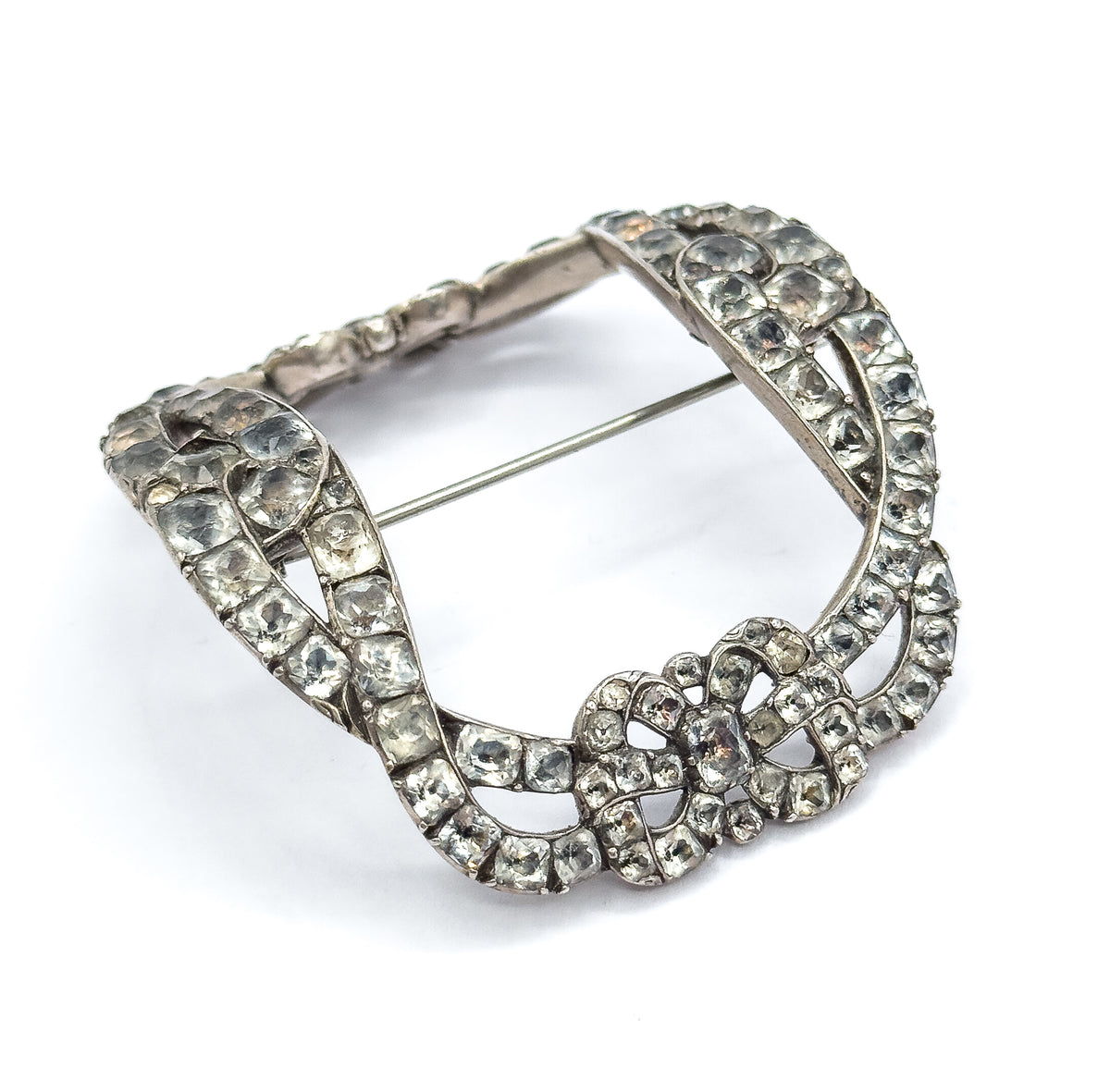 Full three-quarter view of the whole buckle brooch, its interlocking paste-set bands forming an open arched frame around the empty centre, with the pin stem spanning the opening diagonally.