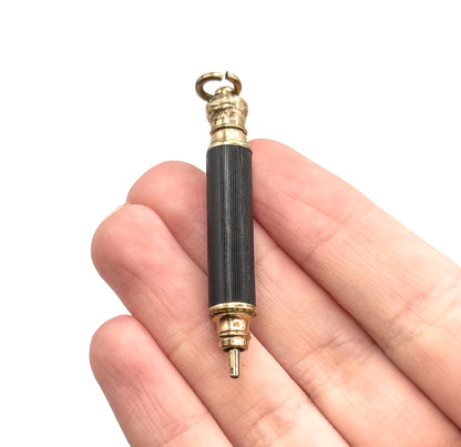 A Victorian mechanical pencil pendant with black textured barrel and gold tone metal ends, shown resting across the fingers of a hand which conveys its small size.