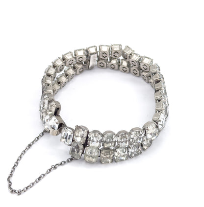 The closed bracelet stands as a ring on white, its fine safety chain hanging loose from the clasp below the two rows of emerald cut glass stones.