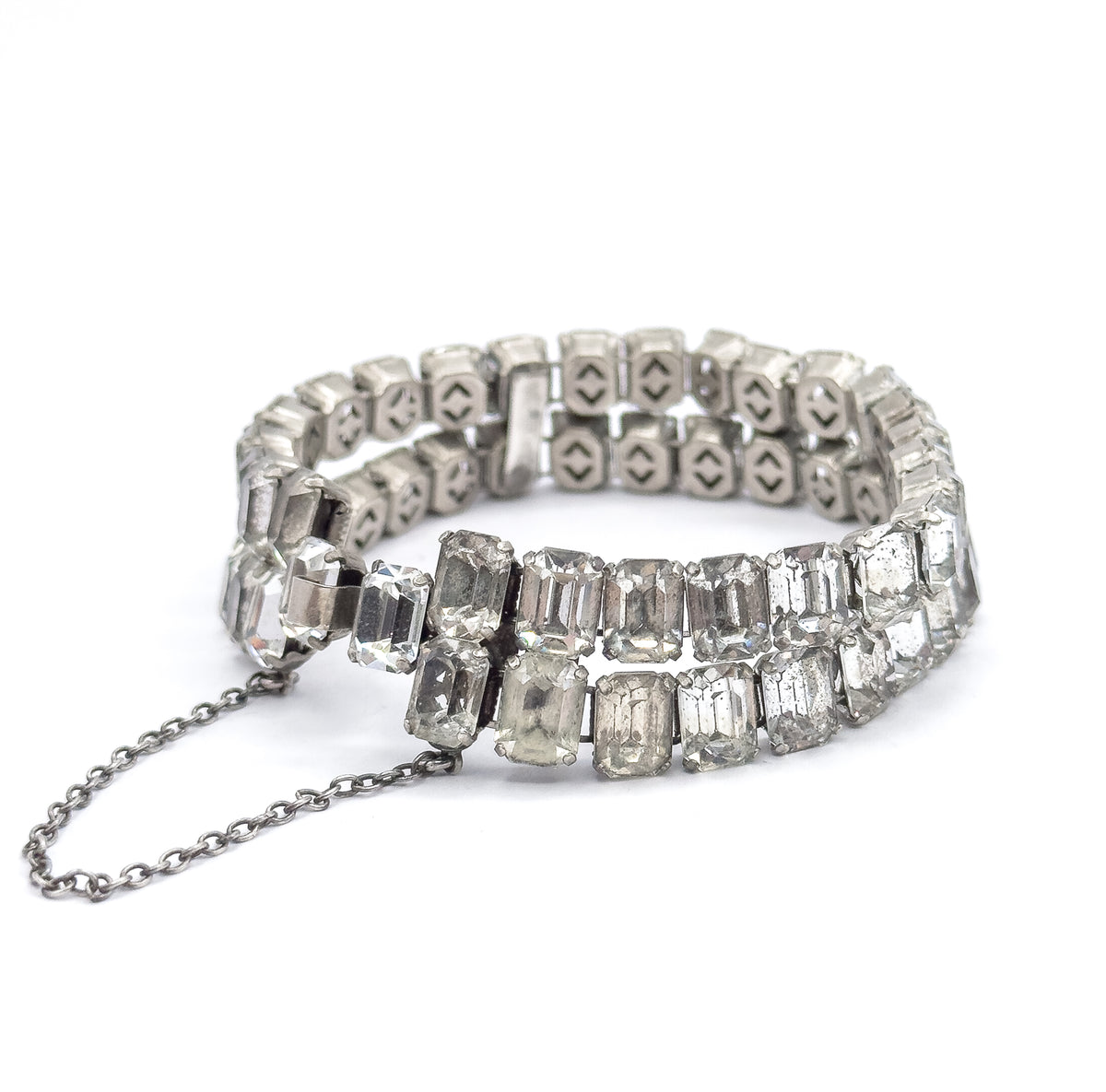 Lower view along the standing bracelet towards the clasp, the safety chain looping onto the white surface and the pierced open backs of the far settings visible above the two rows of glass stones.