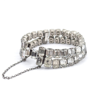 Lower view along the standing bracelet towards the clasp, the safety chain looping onto the white surface and the pierced open backs of the far settings visible above the two rows of glass stones.